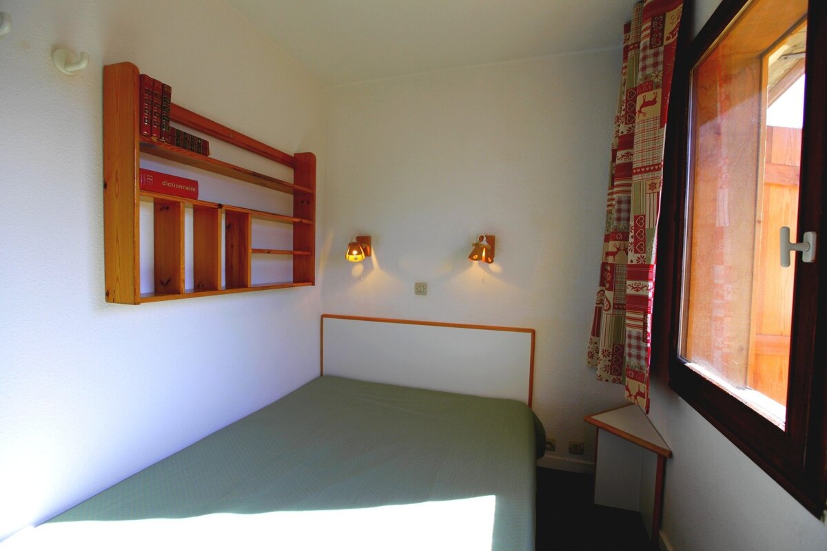 A cozy small bedroom with a single bed, wooden bookshelf, two wall lights, and a window with a red patterned curtain.