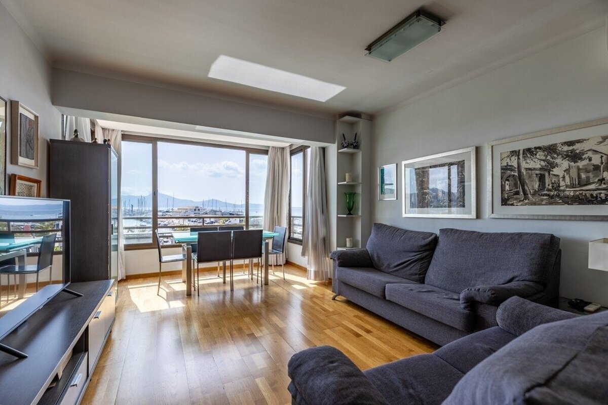 A modern living room with gray sofas, wooden floors, and a dining area next to a large window offering harbor views.