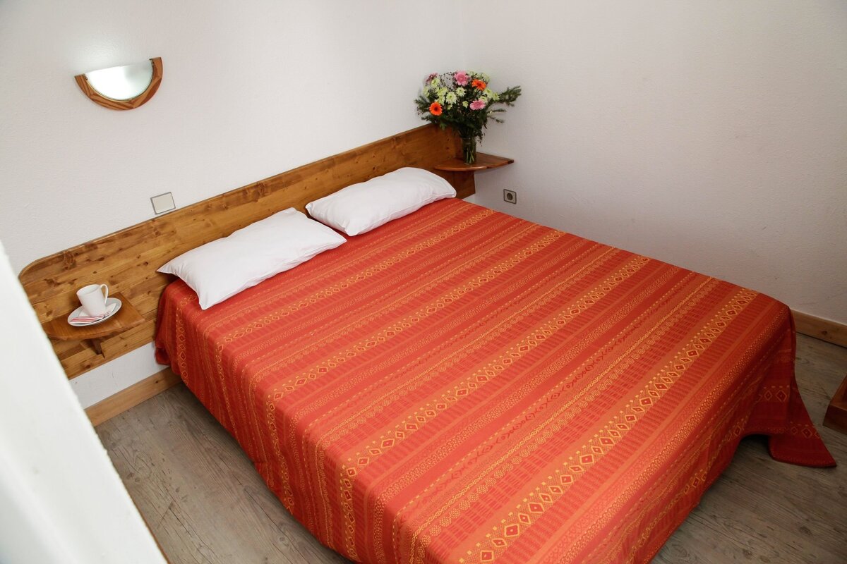 Cozy bedroom with a large bed, vibrant orange patterned bedspread, wooden headboard, white pillows, and a vase of colorful flowers.