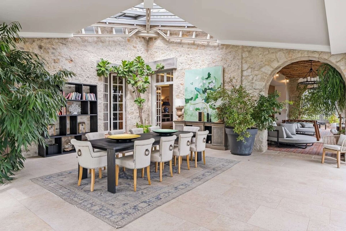 An elegant dining room with stone walls, a skylight, dark table, upholstered chairs, and lush plants. An archway leads to a lounge area.