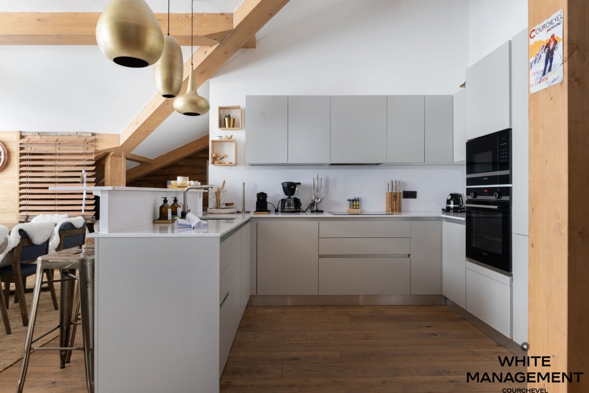A modern, light grey kitchen with sleek cabinets, wooden floors, and exposed beams. Gold pendant lights hang over a counter with a dining area nearby.