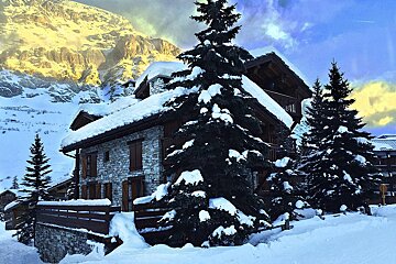 A cozy, snow-covered stone chalet among frosted pine trees. A golden, sunlit mountain peak rises in the background under a vibrant sky.