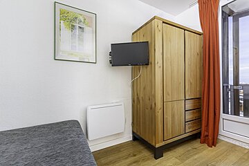 A small room with a wooden wardrobe, wall-mounted TV, heater, framed art, grey seating, and a glass door with an orange curtain.