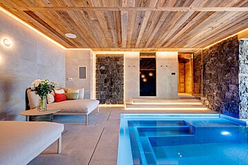 Luxurious indoor spa with a bright blue pool, cozy lounge seating, rustic wooden ceiling, stone accent walls, and warm LED strip lighting, creating a serene ambiance.