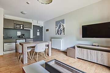 Bright, modern open-plan studio with a kitchenette, light wood dining set, and a living area with a wall-mounted TV. Mountain art and sleek decor.