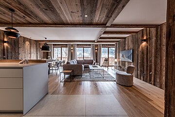 Chic chalet interior: open-plan living/dining, rustic wood walls/ceiling, modern furniture, and large windows revealing a snowy mountain view.