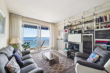 Cozy living room with gray furniture overlooking a bright blue ocean from a balcony with blue awnings. Shelves are filled with books and decor.