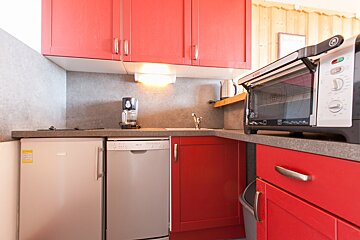 A compact kitchen featuring vibrant red cabinets, grey countertops, and appliances including a fridge, dishwasher, coffee maker, kettle, and oven.