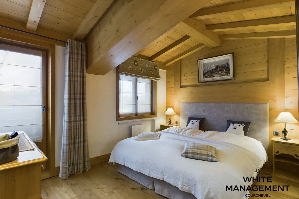 A warm, rustic bedroom featuring extensive wood paneling, exposed beams, a spacious bed with polar bear pillows, and natural light from windows.