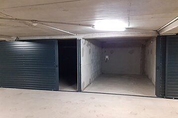 A brightly lit underground garage showing two empty concrete storage units. One dark blue sliding door is partially open, the other is mostly closed.