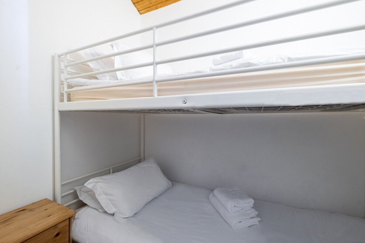 A clean, minimalist room features white metal bunk beds with fresh white bedding and folded towels. A wooden bedside table is on the left, with a wooden ceiling above.