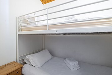 A clean, minimalist room features white metal bunk beds with fresh white bedding and folded towels. A wooden bedside table is on the left, with a wooden ceiling above.