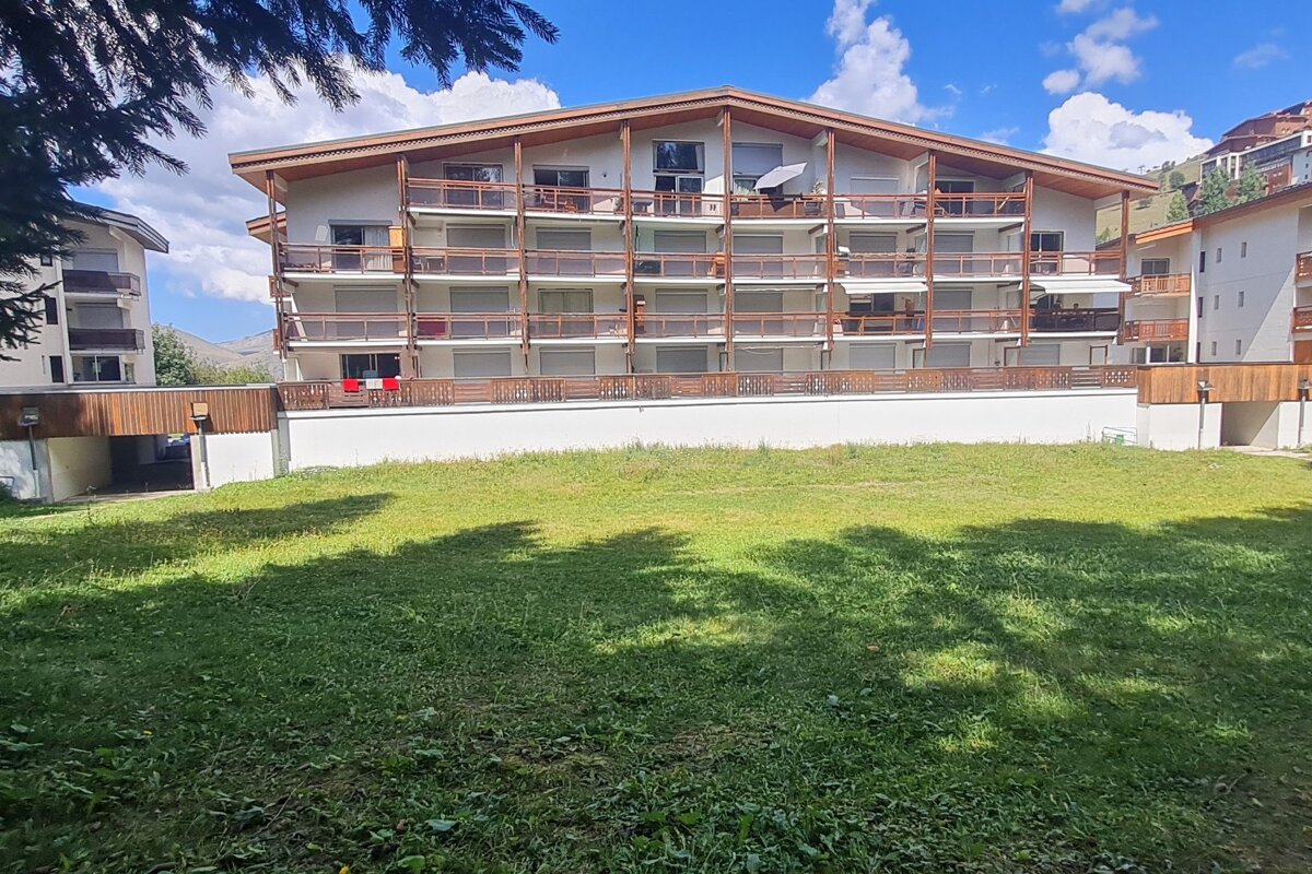 A multi-story alpine-style building with balconies overlooks a green lawn under a sunny, cloudy sky. Distant mountains are visible.