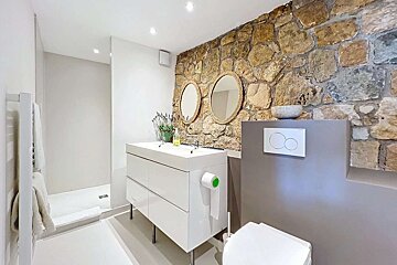 Modern bathroom featuring a rustic stone wall, white double vanity, two round mirrors, and a sleek shower area.