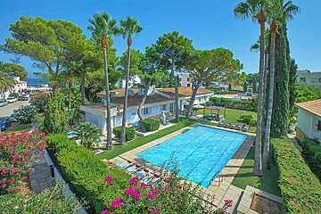 A vibrant resort features a large blue swimming pool, lush green lawns, a white building, and tall palm trees under a clear blue sky, with pink flowers.