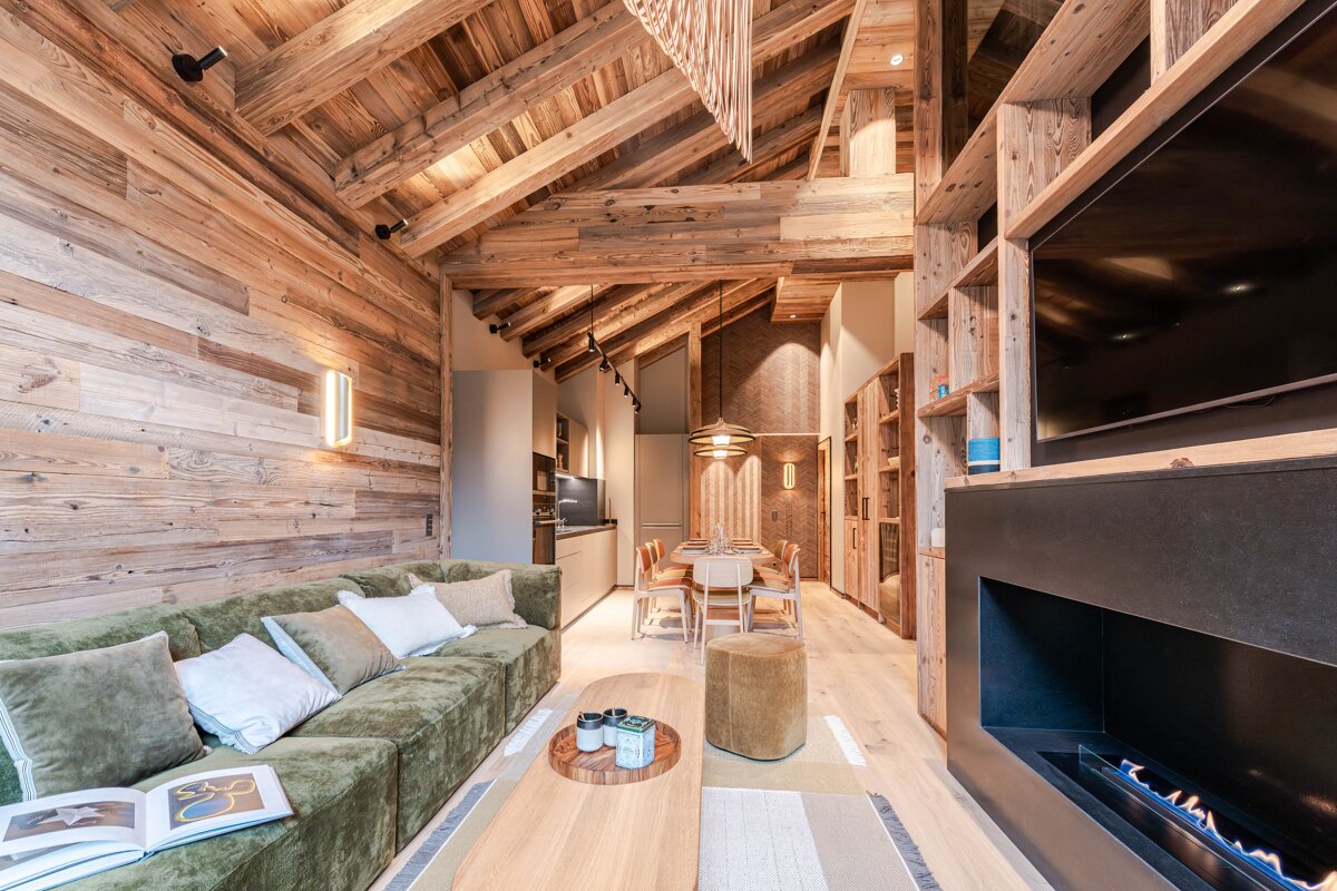 A rustic modern chalet living area with wood walls/ceiling, green sofa, fireplace, TV, and an open dining/kitchen space.