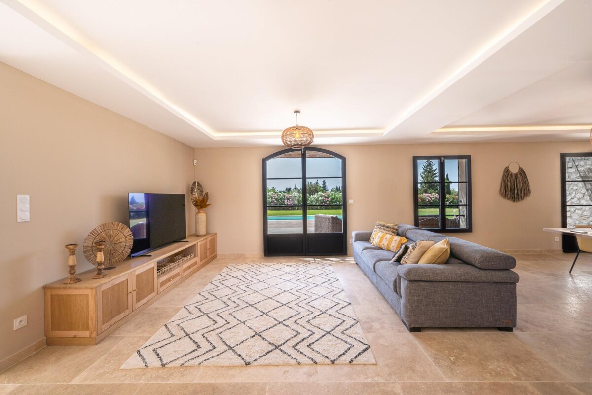 A stylish living room with a gray sofa, TV, patterned rug, and arched doors revealing a pool and garden. Neutral tones and soft lighting.