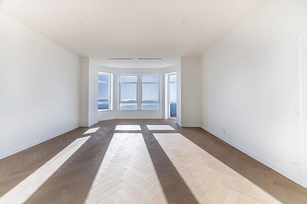 Bright, empty room with white walls, herringbone wood flooring, and windows offering a scenic view.