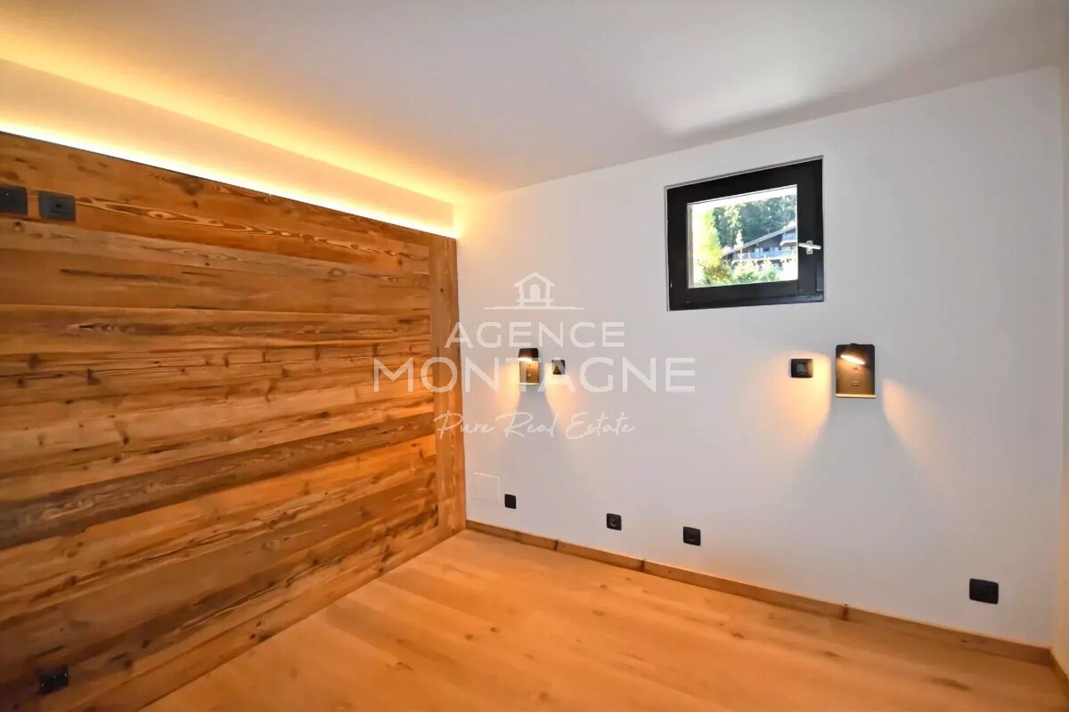 Cozy room featuring a backlit wooden accent wall, light wood flooring, white walls with sconces, and a small high window.