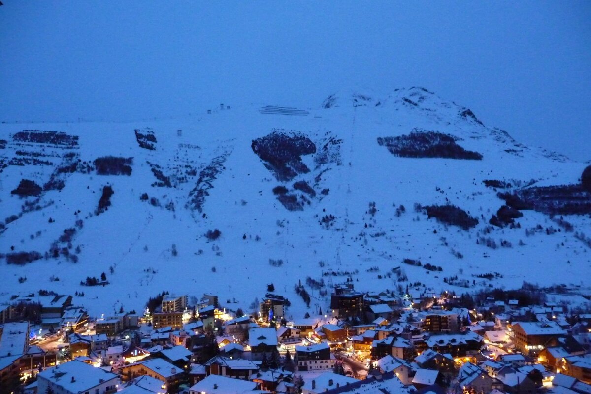 A snow-covered mountain town at dusk. Warm lights from buildings glow under a dark blue sky and imposing snowy peaks.