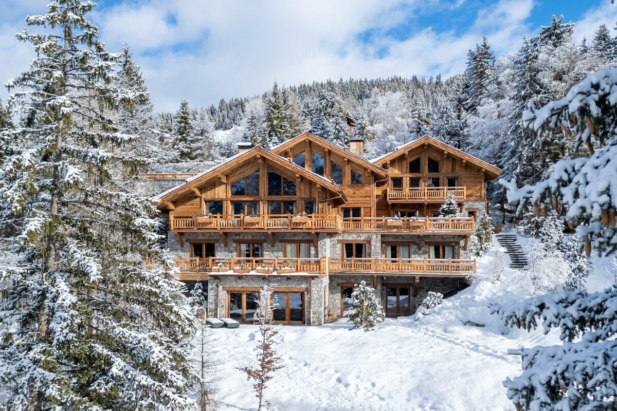 A grand wooden and stone chalet blanketed in snow, nestled among snow-covered pine trees and mountains under a clear blue sky.