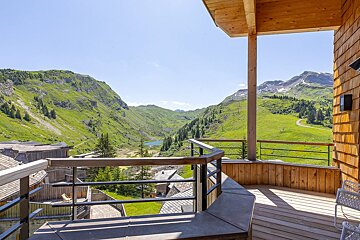View from a wooden balcony overlooking a lush green mountain valley and lake under a clear blue sky.
