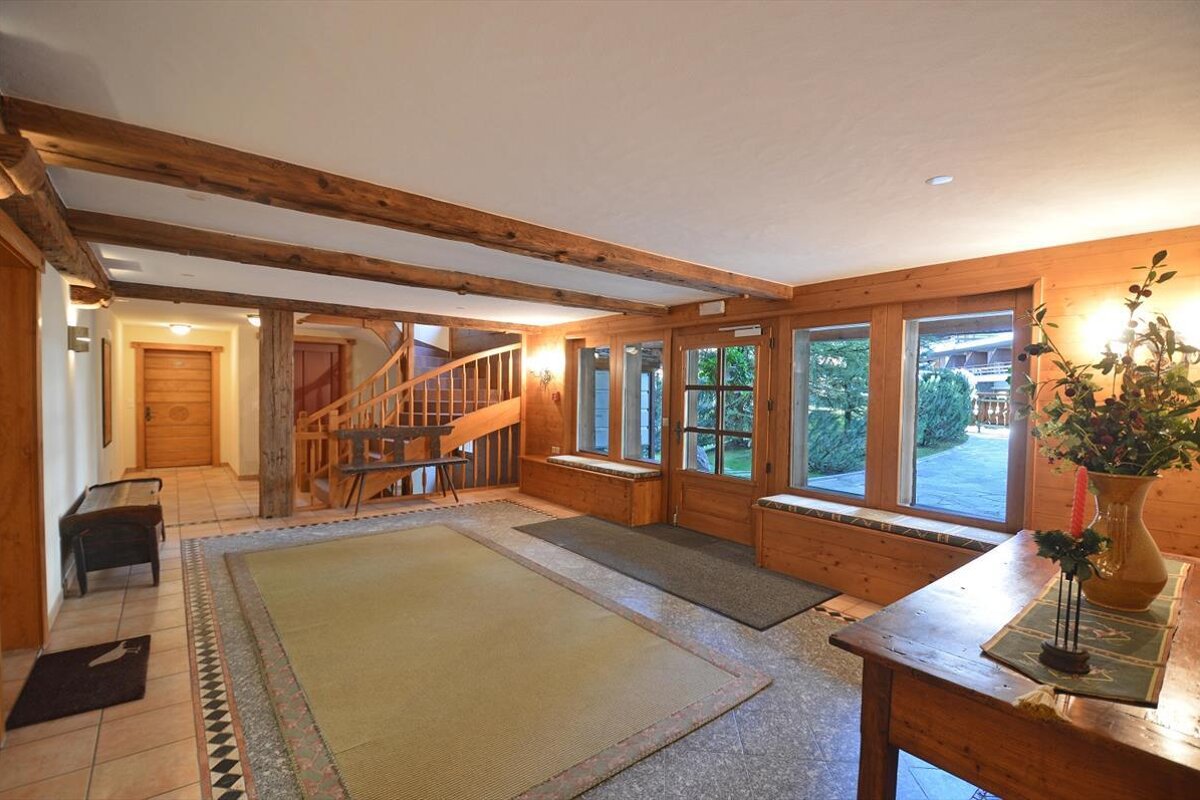 A warm, rustic hall with exposed wooden beams, a curved staircase, large windows, and tiled floors with rugs, creating a cozy atmosphere.