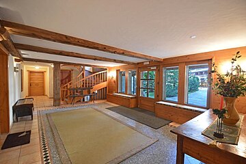 A warm, rustic hall with exposed wooden beams, a curved staircase, large windows, and tiled floors with rugs, creating a cozy atmosphere.