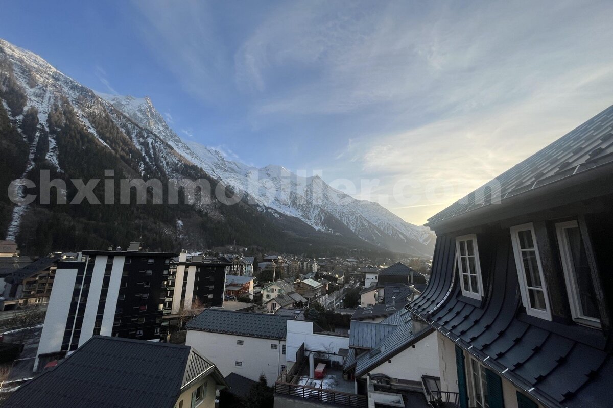 Snowy mountain range overlooking a valley town, with building rooftops in the foreground under a soft, clear sky.
