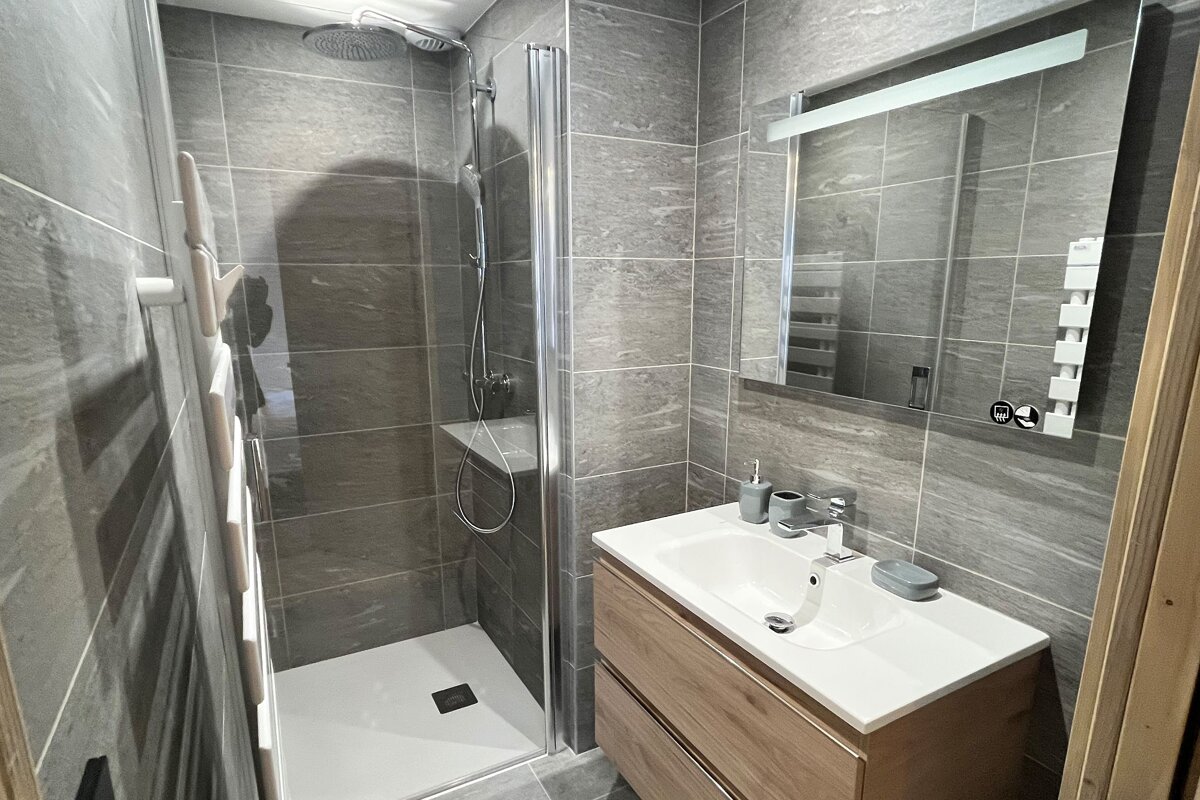 A modern bathroom features gray tiled walls, a glass-enclosed shower, a vanity with a white sink, and a lighted mirror. A white towel warmer is on the left.