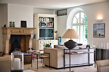 A bright, elegant living room with a stone fireplace, bookshelf with TV, sofa, and a large arched window, featuring neutral decor.