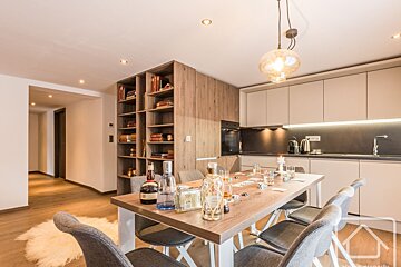 An elegant dining room with a wooden table, chairs, shelving unit, and a small kitchen in the background.