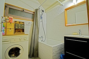 A compact, white-tiled bathroom with a washing machine, bathtub with shower, vanity sink, and mirror. A small window is above the washer.