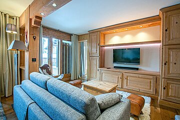 Cozy chalet living room featuring wooden walls, a grey sofa, and a built-in entertainment unit with a TV. Large glass doors lead outside.