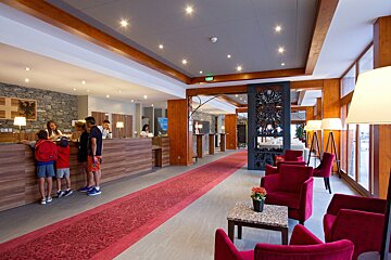 A welcoming hotel lobby with a wood-paneled reception desk where a family checks in, a long red carpet, and a cozy lounge area with red velvet armchairs.