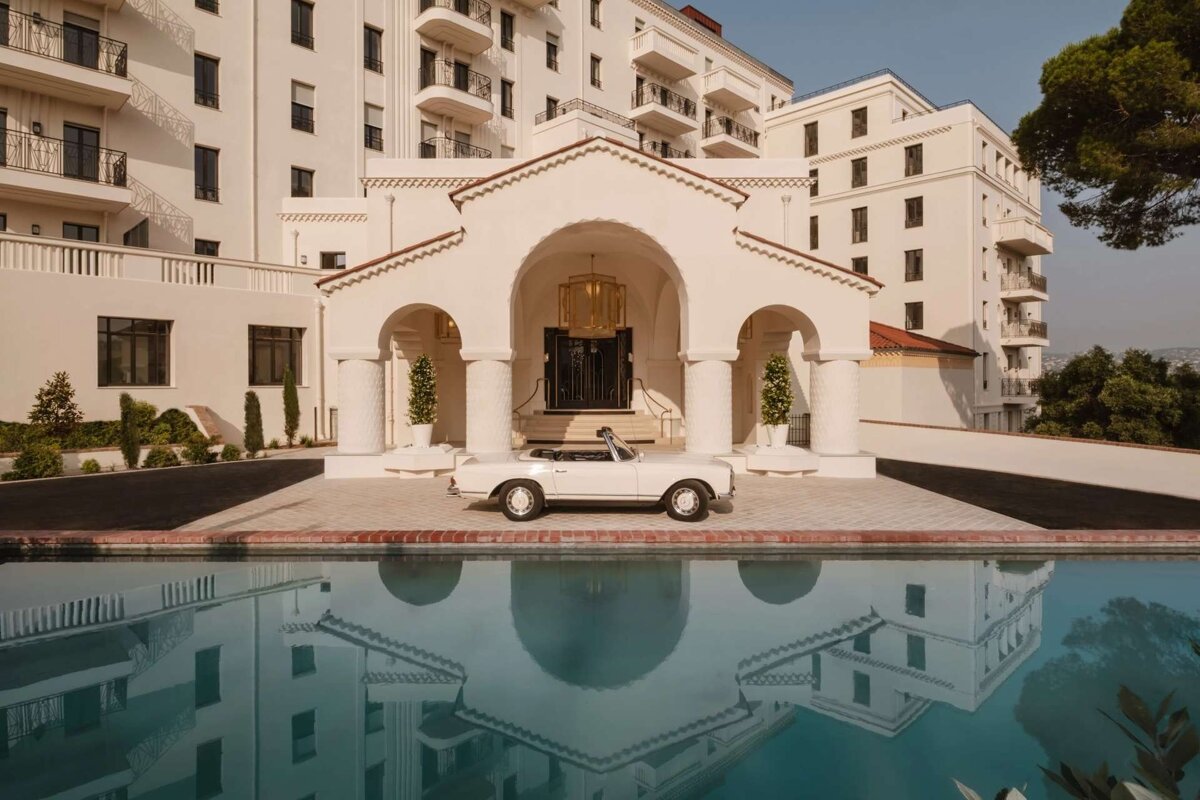 An elegant white building with an arched entrance, a vintage white convertible, and its reflection in a still pool under a bright sky.