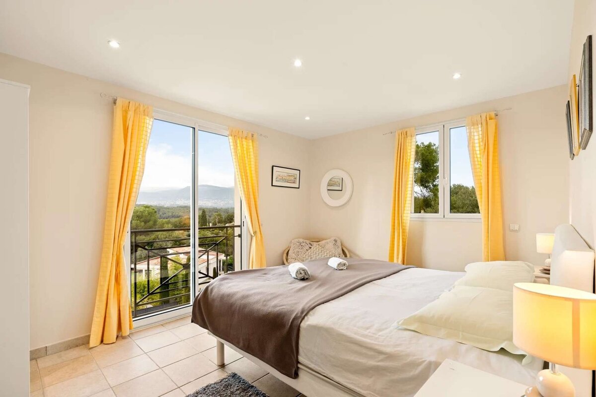 Bright, modern bedroom with a comfortable bed, vibrant yellow curtains, and a balcony offering a scenic view of mountains, trees, and houses.