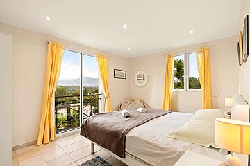 Bright, modern bedroom with a comfortable bed, vibrant yellow curtains, and a balcony offering a scenic view of mountains, trees, and houses.