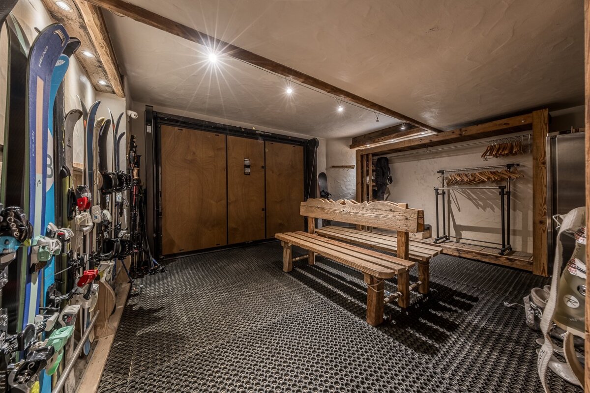 A rustic ski room with skis, wooden benches, and storage lockers. Textured floor, exposed beams, and warm lighting.