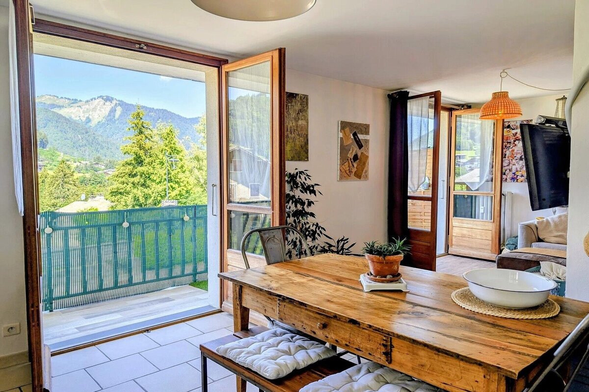 A bright dining room with a rustic wooden table, opening onto a balcony with scenic mountain views.