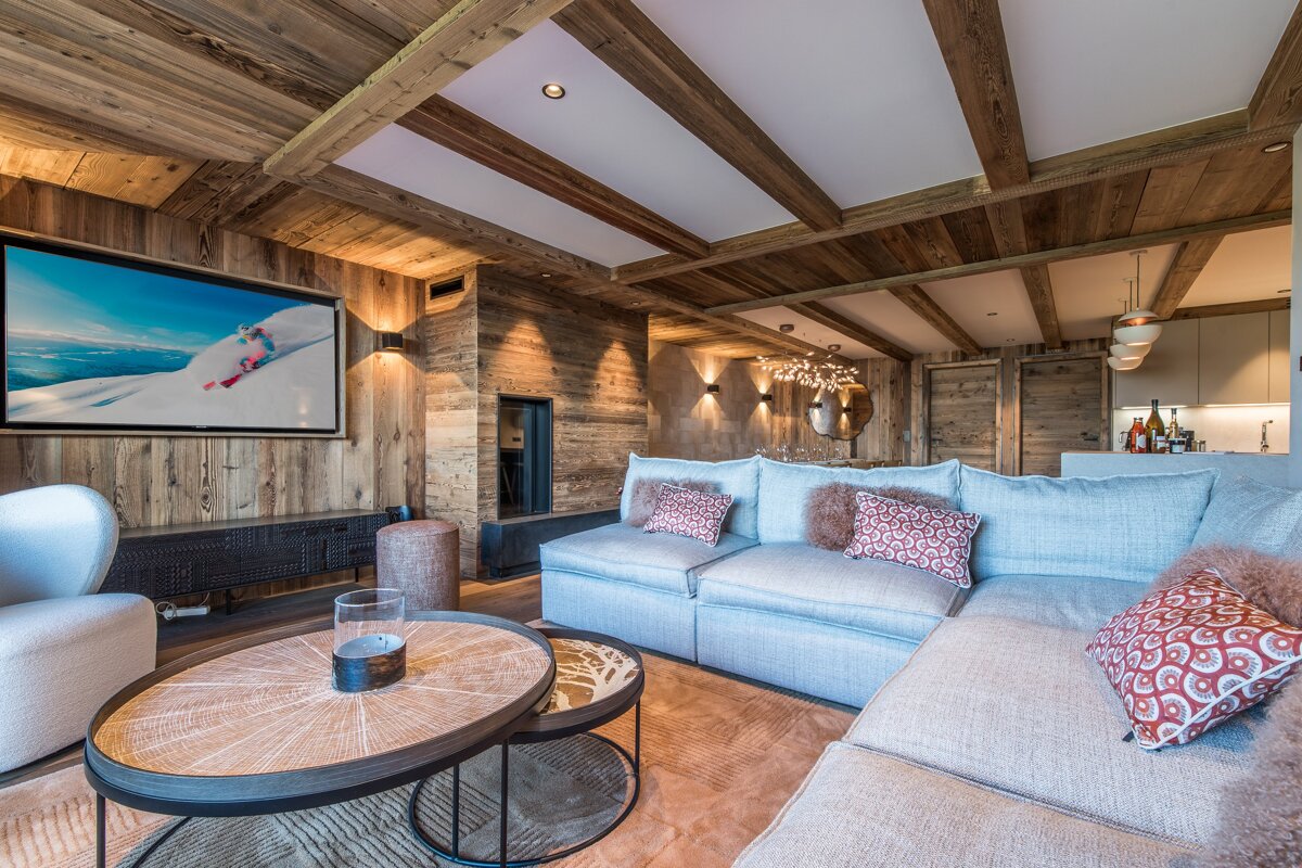 A rustic modern living room with wood walls/ceiling, a large light blue sectional sofa, a TV showing skiing, and a modern fireplace.