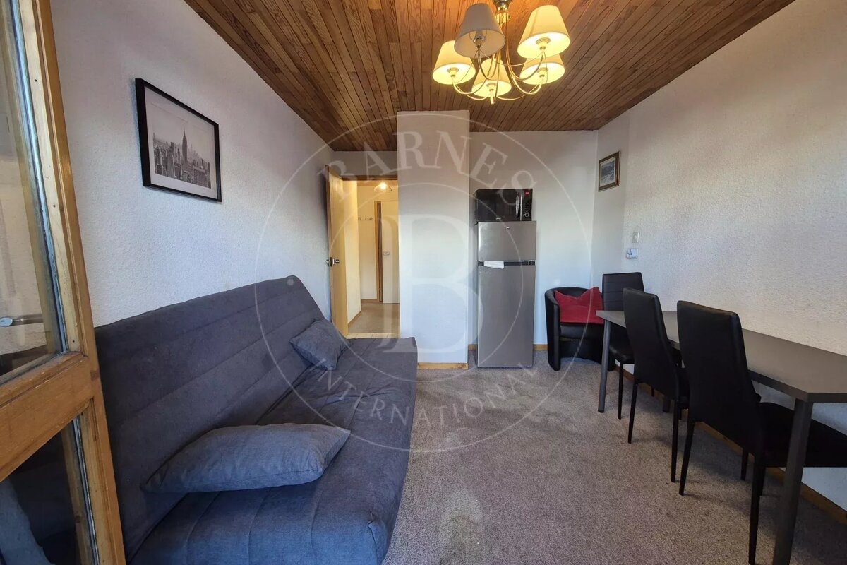 A cozy, compact living/dining area with a grey sofa, dining set, and kitchen appliances, featuring a warm wooden ceiling and white walls.
