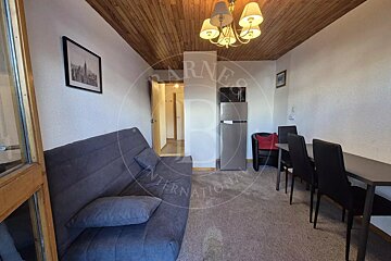 Small living area with a grey sofa, wood-paneled ceiling, dining table with black chairs, and a refrigerator in the background.