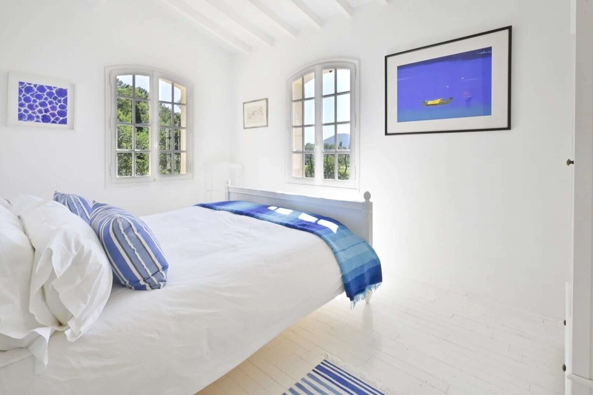 Bright, minimalist white bedroom with blue accents on bedding, a striped rug, and wall art. Two arched windows frame green outdoor views.