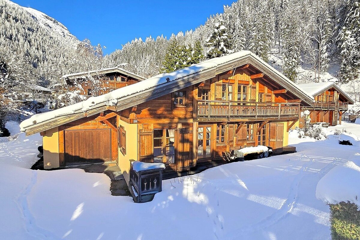 Snow-covered wooden chalet on a sunny winter day, surrounded by pine forests and mountains. Deep snow blankets the ground and roofs.