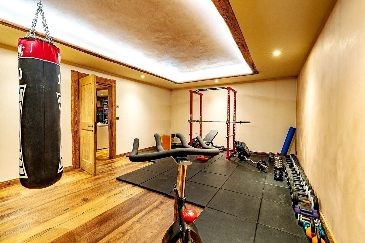 Modern home gym with a punching bag, exercise bike, power rack, and dumbbells. Features warm lighting, wood floors, and rubber mats.