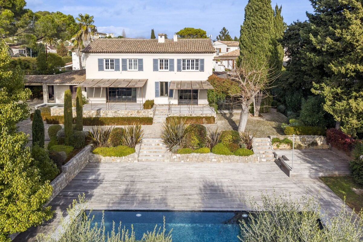 A luxury two-story villa with blue shutters, landscaped gardens, a spacious wooden deck, and a swimming pool.