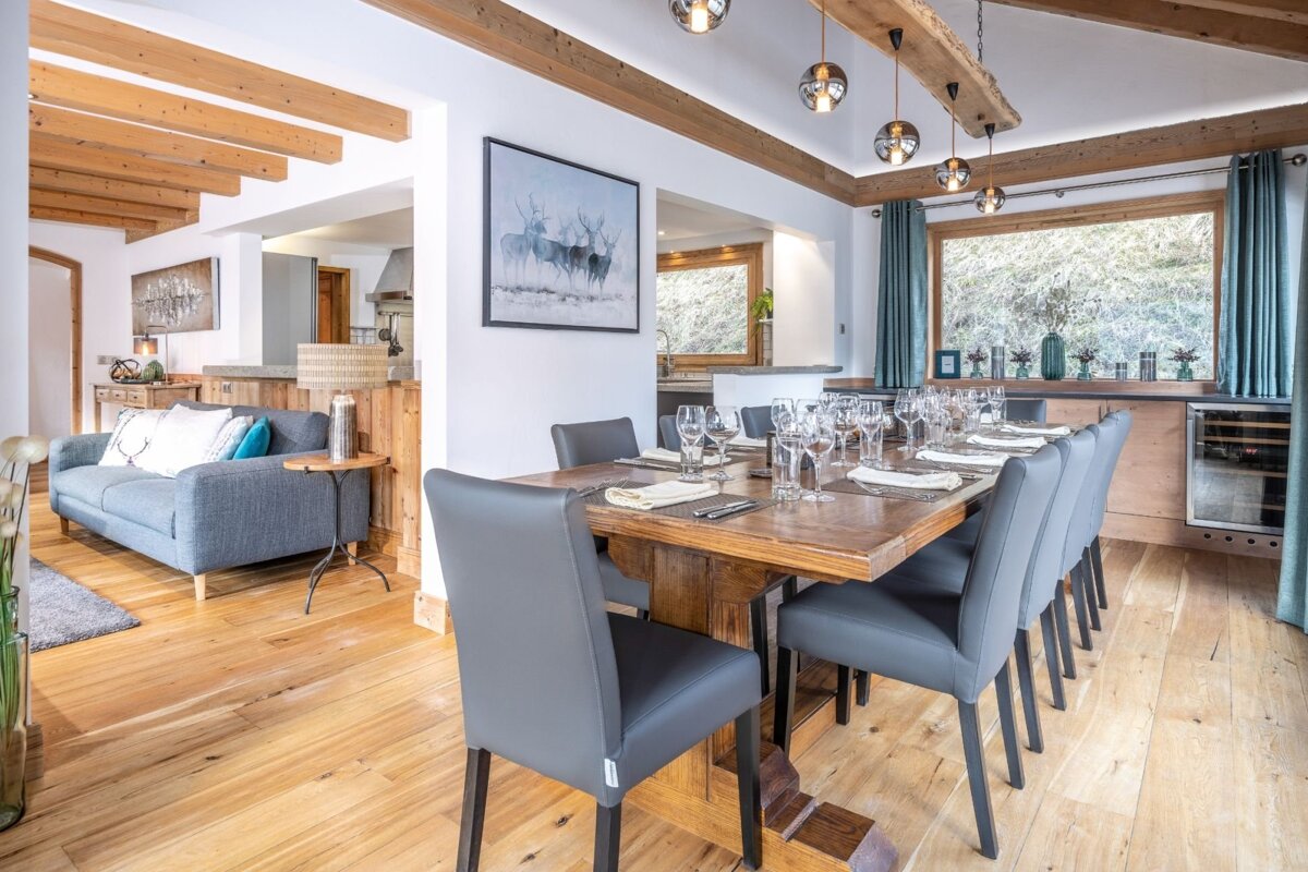 A cozy, open-plan living and dining room with rustic wood beams, a long dining table set for guests, and a sofa area nearby.