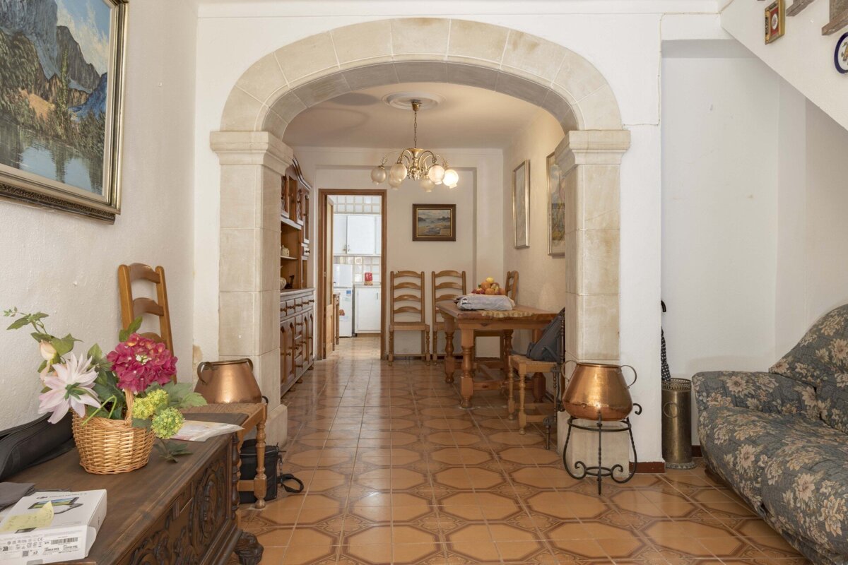 A cozy interior features a large stone archway separating a living area with a floral sofa from a dining space with a wooden table, leading to a kitchen.
