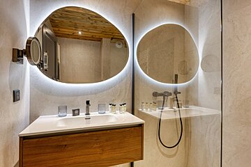 A stylish modern bathroom with organic-shaped, backlit mirrors, a wooden vanity, and a glass shower. Warm neutral tones throughout.